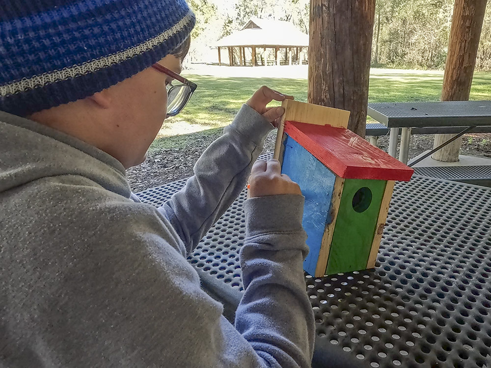 boy wearing grey sweatshirt, blue cap and glasses paints a wood birdhouse