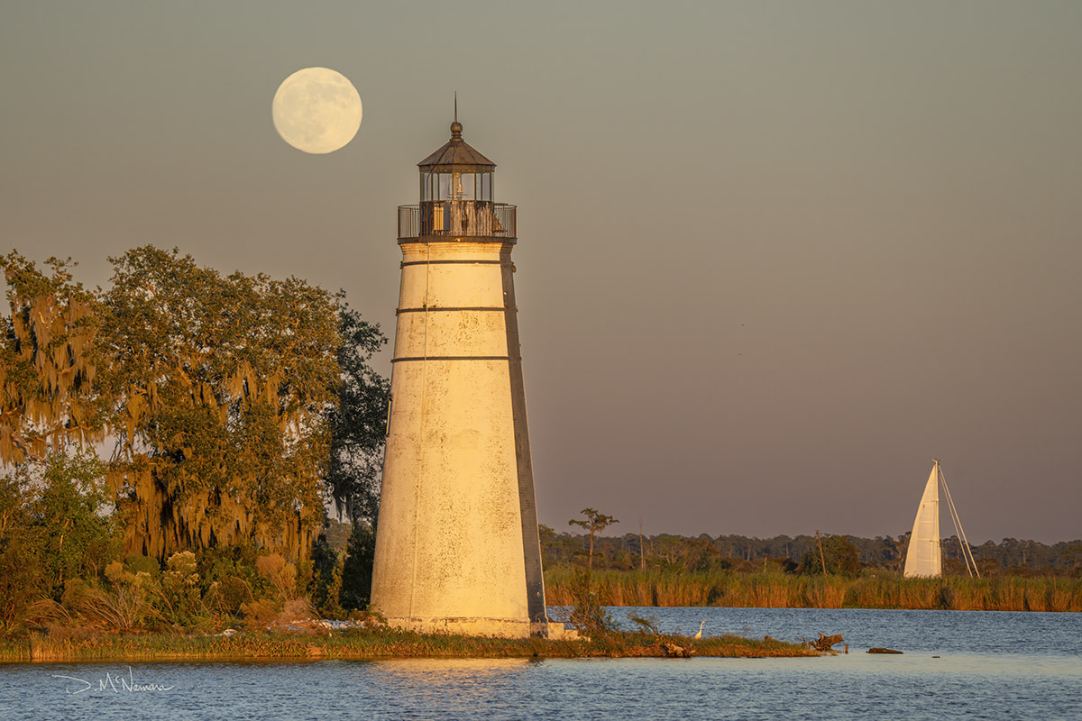 full moon near lighthouse on lake with sailboat in the background at sunset