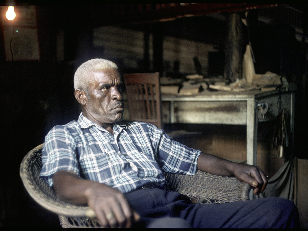 photograph of black man with gray hair sitting in chair wearing plaid shirt