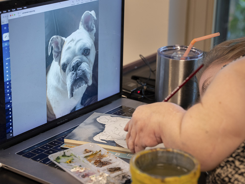 artist lies next to computer with image of white dog as she paints