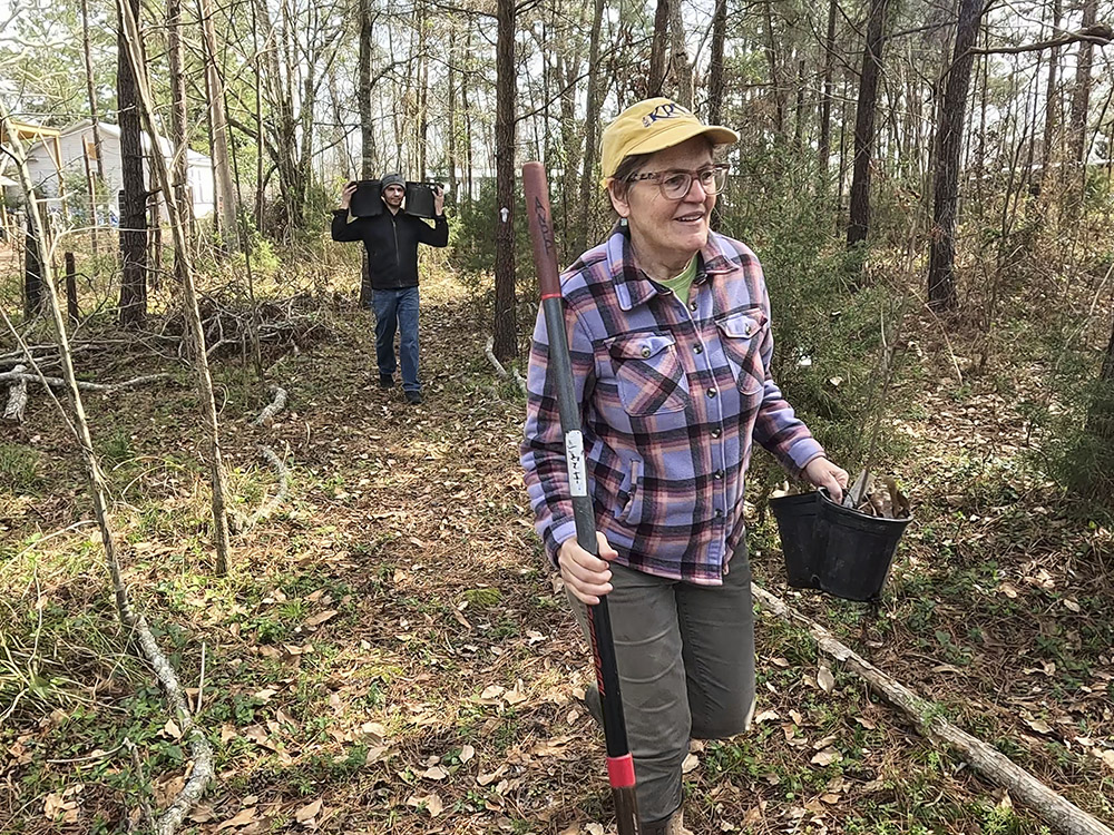 woman in yellow cap and plaid shirt carrying shovel follwed by young man dressed in black carring potted plants