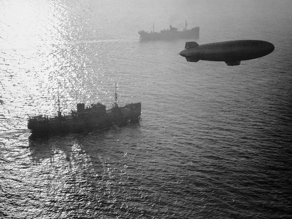 blimp flies above ships in black and white photograph