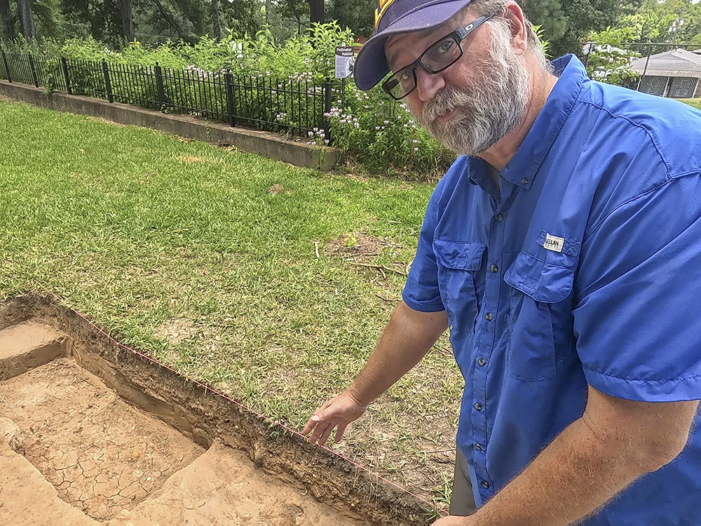 man in cap with beard and glasses points to square hole in earth
