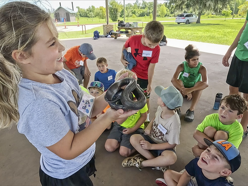 young girl in light bluie shirt holds black snake in front of other children
