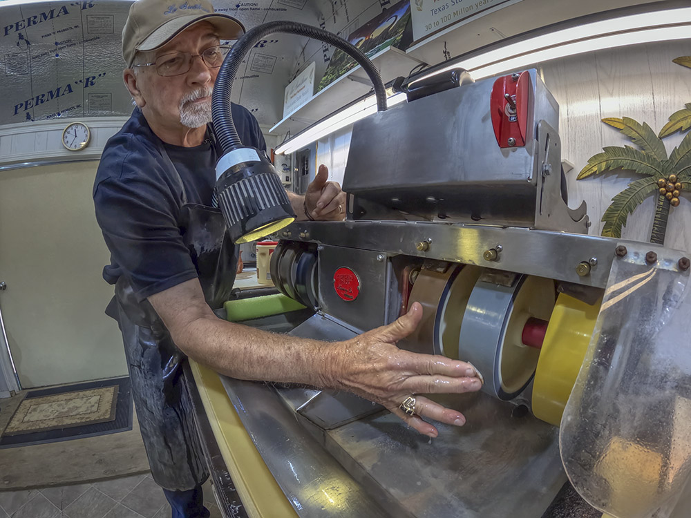 man wearing cap, dark shirt and apron polishes small stone on machine