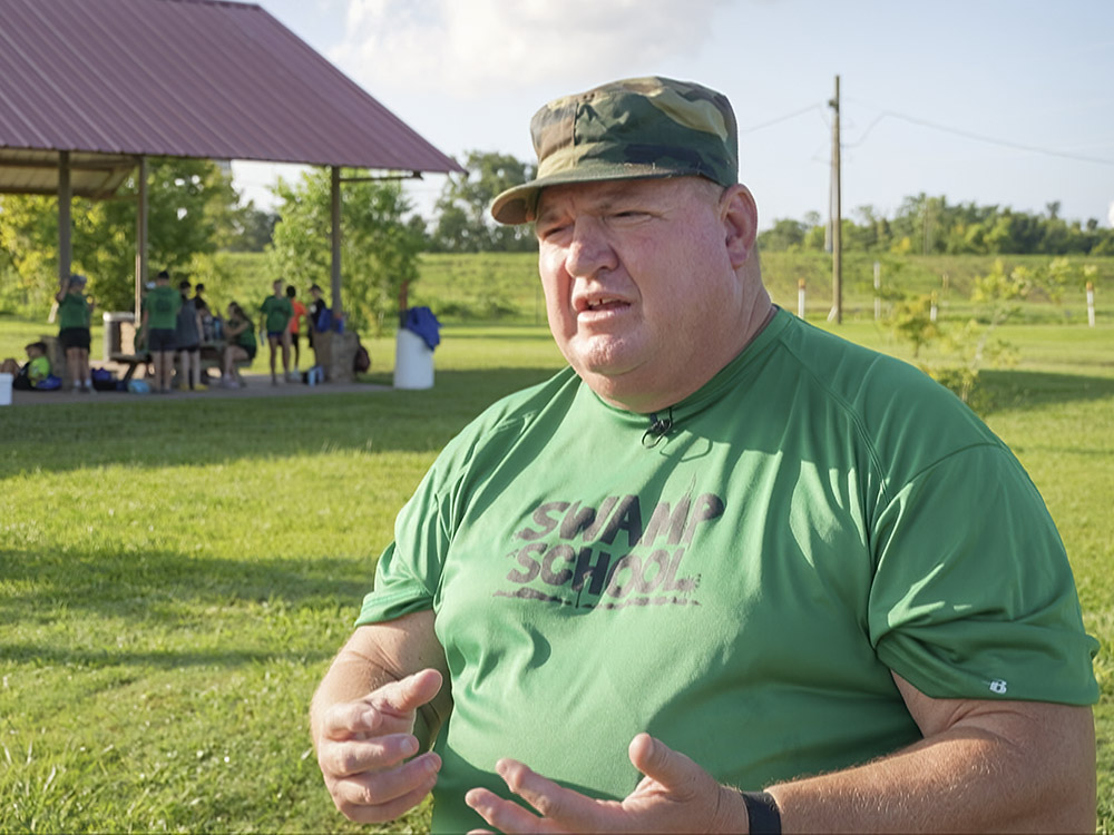 man wearing green swamp school t-shirt and camo hat talks outdoors