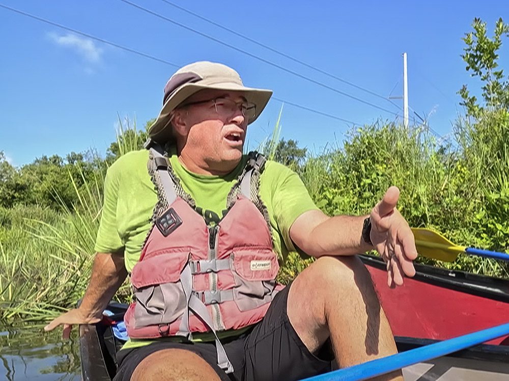 man wearing life jacket, green shirt and hat talking to class in canoe in swamp school