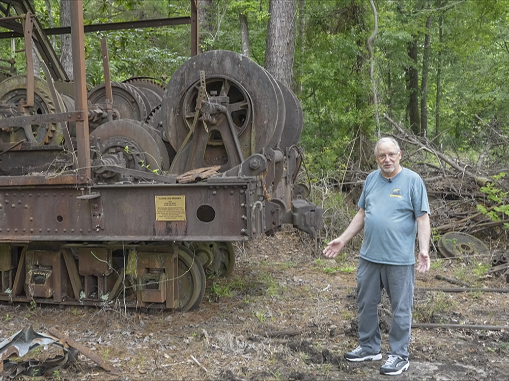 man with gray hair, blue shirt and jeans stands next to rusted rail equipment