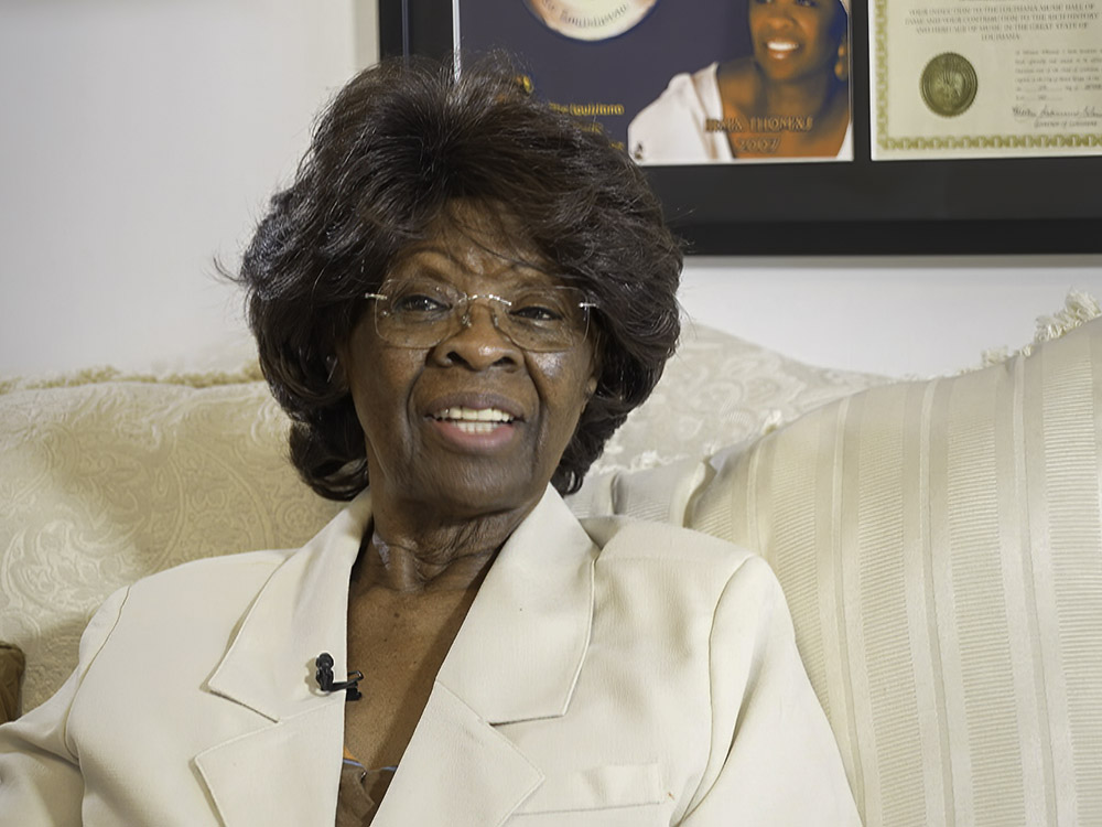 woman with glasses, dark hair and white suit jacket sits on white sofa