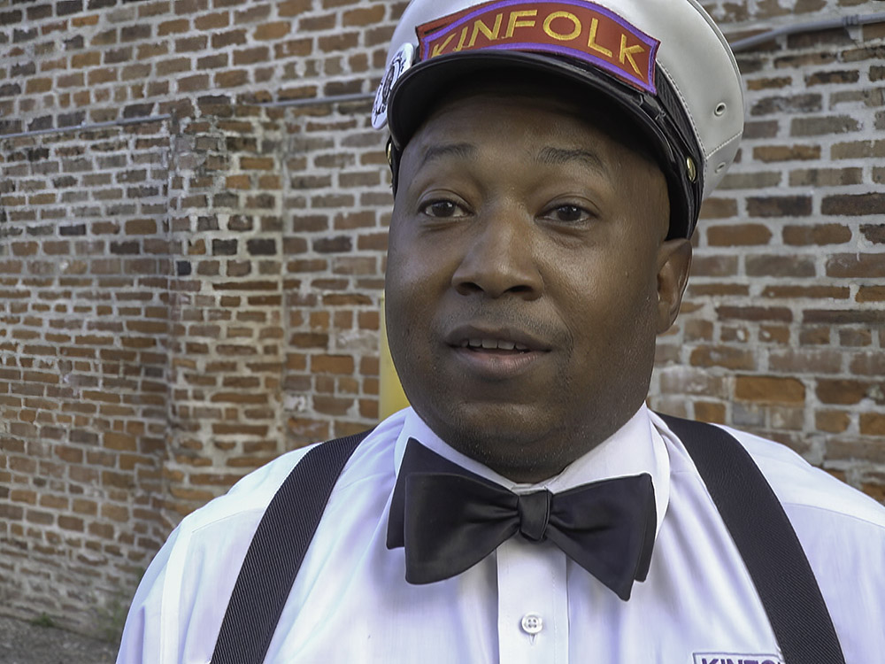 man wearing cap with word kinfolk, white shirt with black suspenders and bow tie