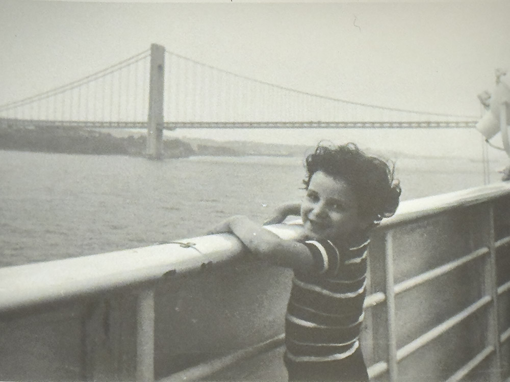 black and white photo of young boy at ship's railing near suspension bridge
