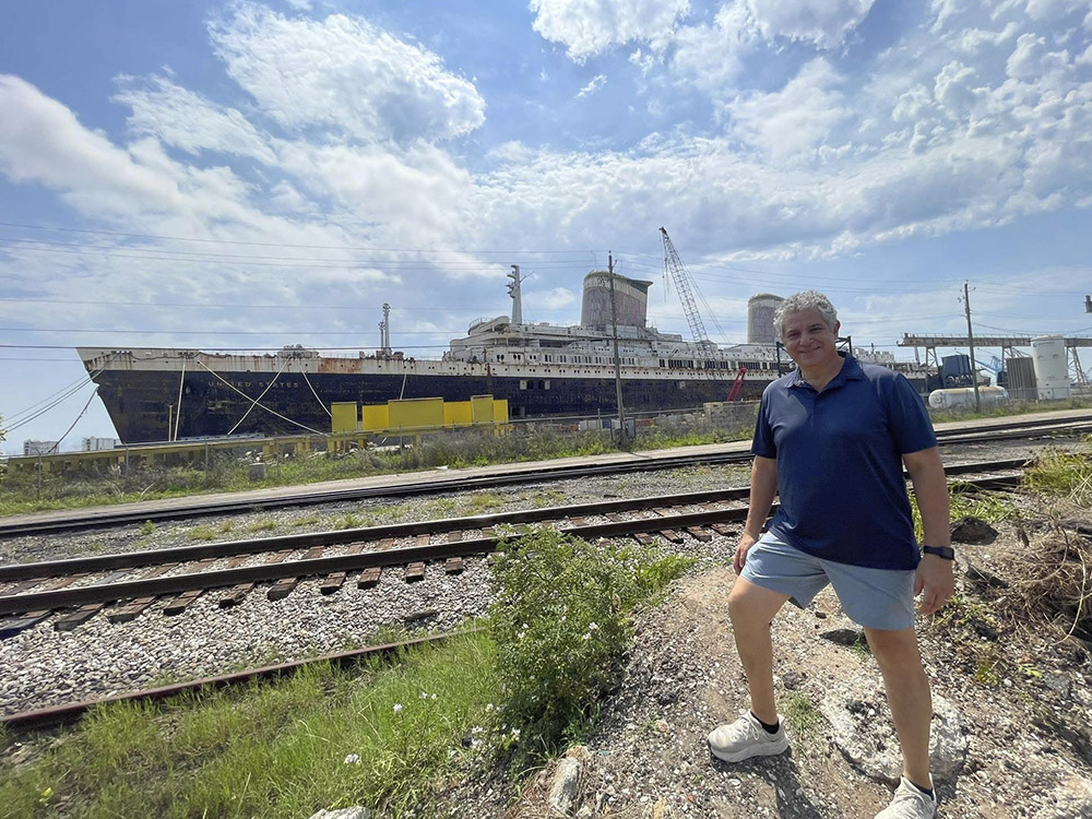 man with dark blue shirt and light blue shorts standing in front of SS United States in port