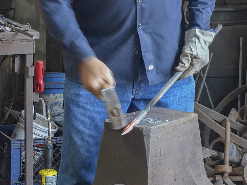man in blue work clothes strikes piece of hot steel with hammer on anvil