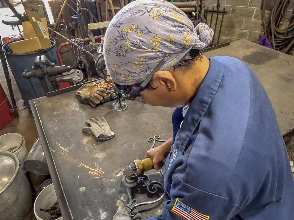 woman wearing bandana on head, workglasses and blue shirt polishes ironwork
