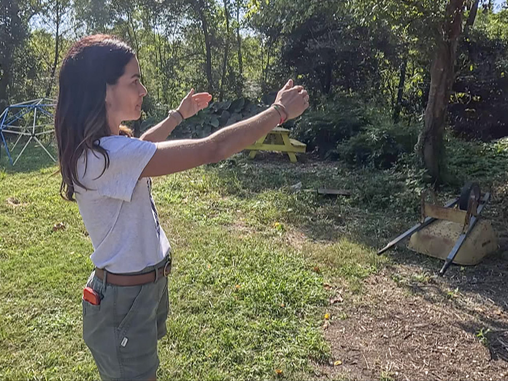 woman with dark hair, light gray shird and dark gray shorts standing in field gesturing with arms