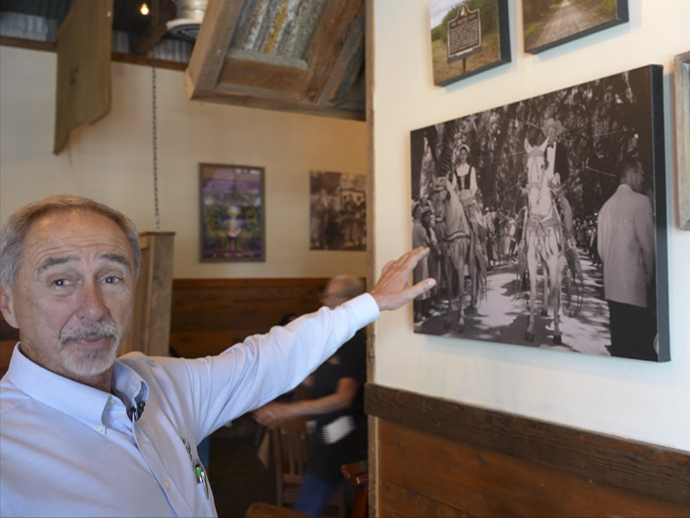 man in longsleeve blue shirt points to black and white photo on wall of people on horseback