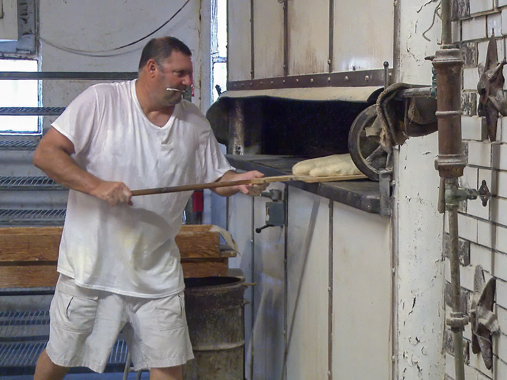 man with short hair, white t-shirt and white shorts uses paddle to put bread in oven