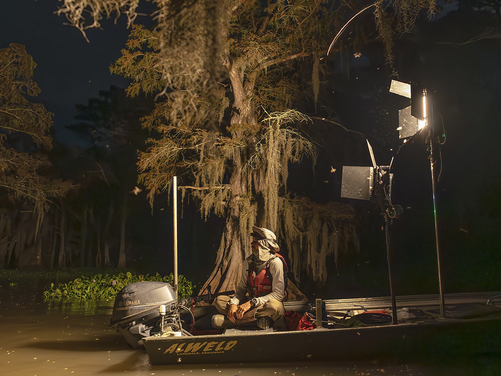 man in hat sitting in flatboat with studio lights in cypress swamp