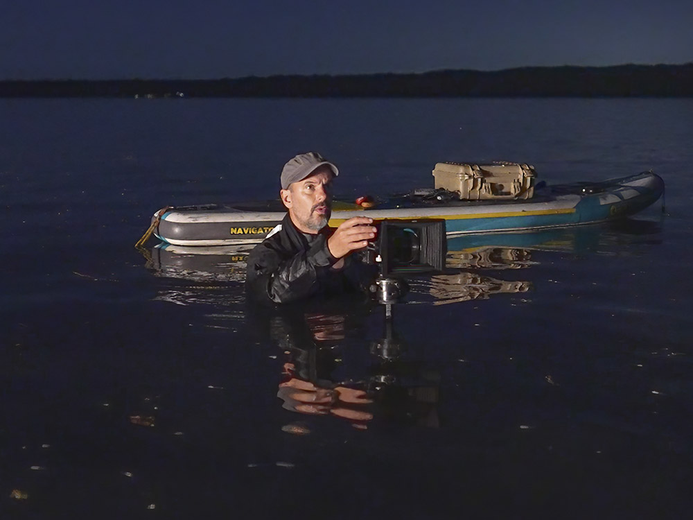 man in dark clothing and cap standing in chest deep water with camera on tripod near paddle board