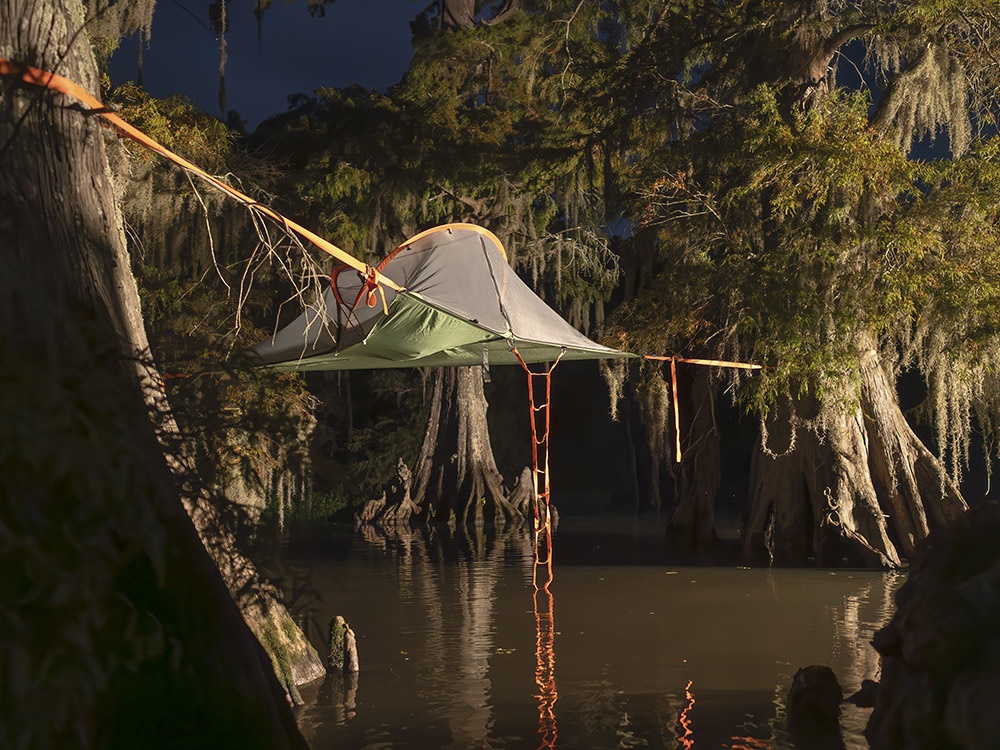 a hammock tent suspended between cypress trees hanging above the water at night