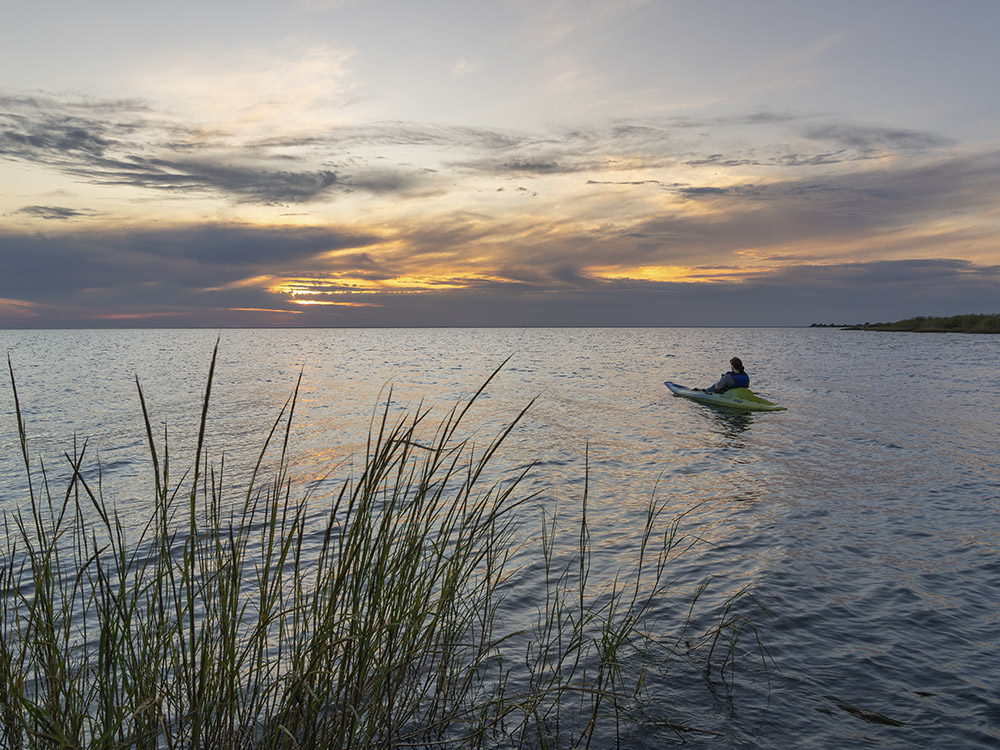 person in kayak watches sunset behind clouds over lake