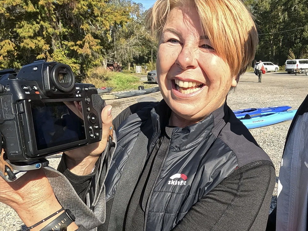 woman with short blond hair wearing black jacket and holding camera