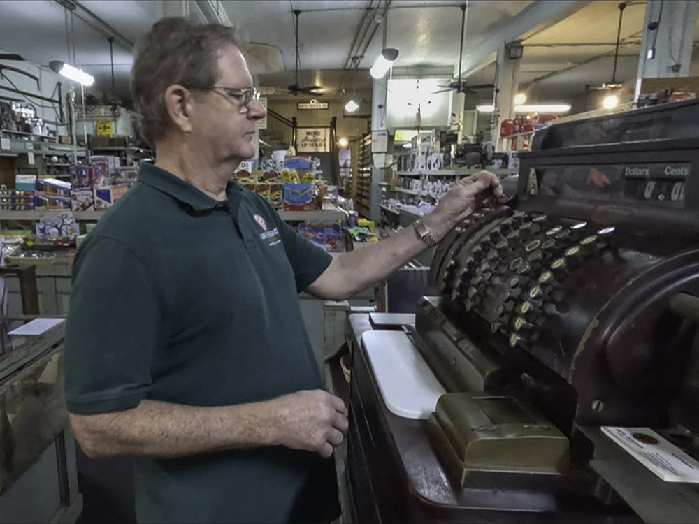 man wearing dark green shirt and glasses operates an antique cash register