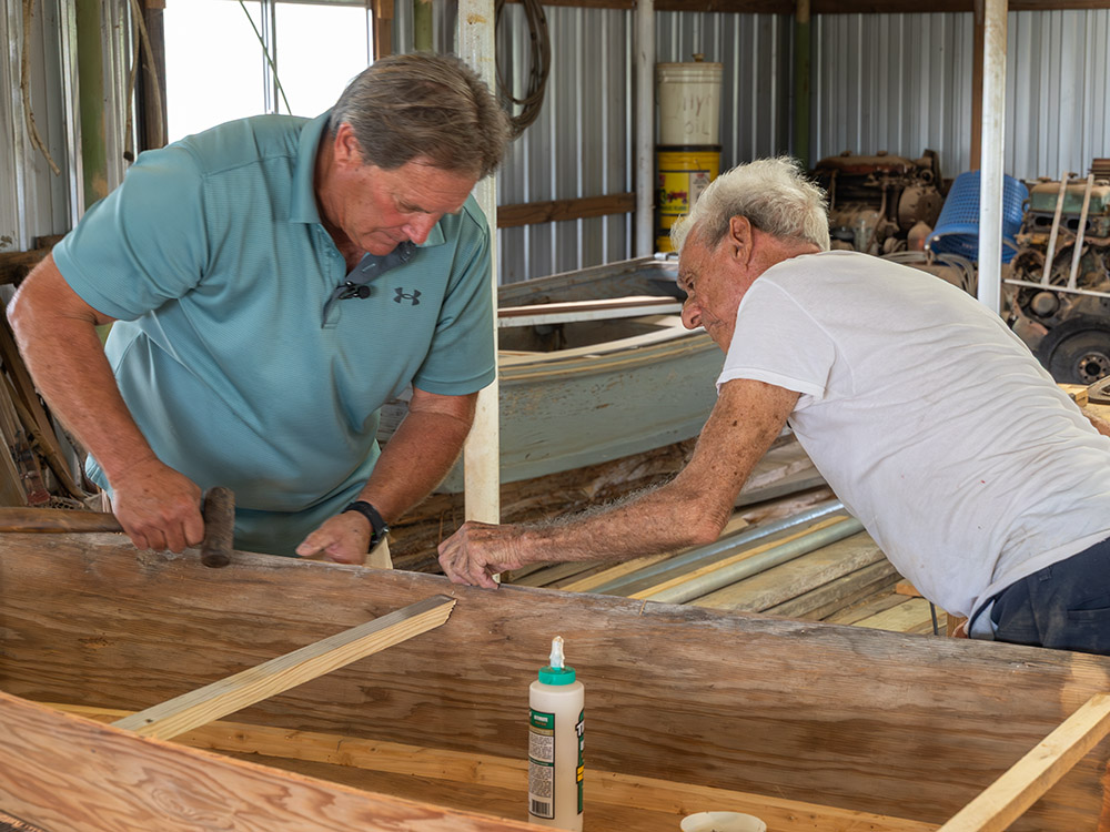 man in green shirt works with elderly man wearing white t-shirt building small wood boat