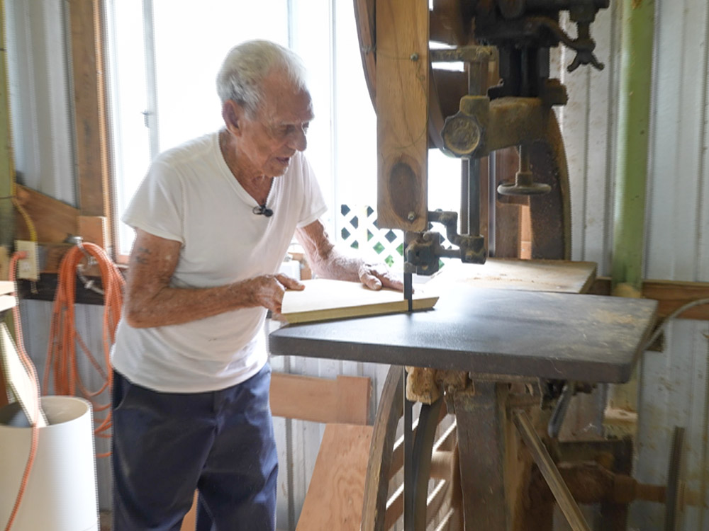 elderly man wearing white t-shirt and blue jeans cuts wood on band saw