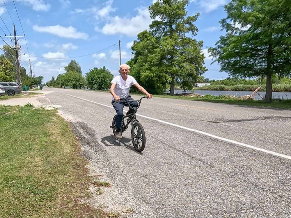 elderly man wearing jeans and white t-shirt rides bicycle along side of highway