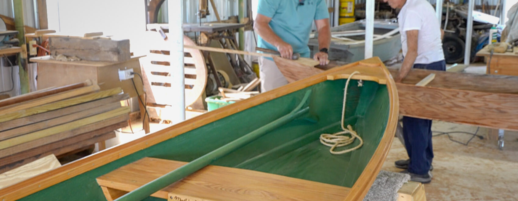 two men work to build boat in workshop[ with green pirogue in foreground