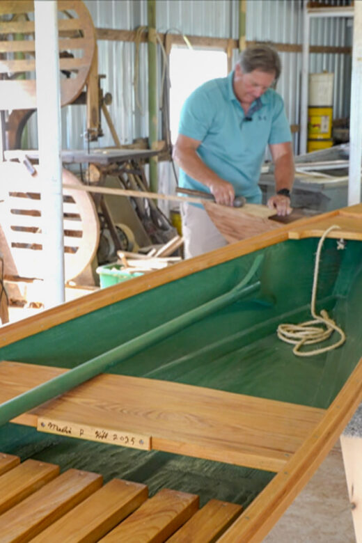 two men work to build boat in workshop[ with green pirogue in foreground