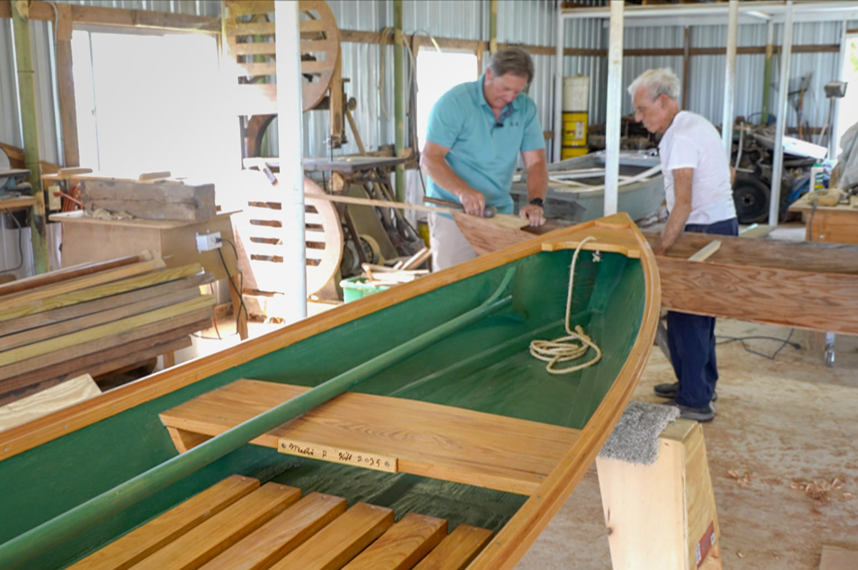 two men work to build boat in workshop[ with green pirogue in foreground